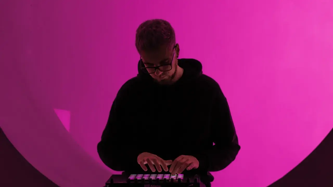 Un homme en sweat à capuche noir joue sur un contrôleur Ableton, illuminé par une lumière rose. Il est plongé dans sa création, un moment clé de la culture musicale moderne.