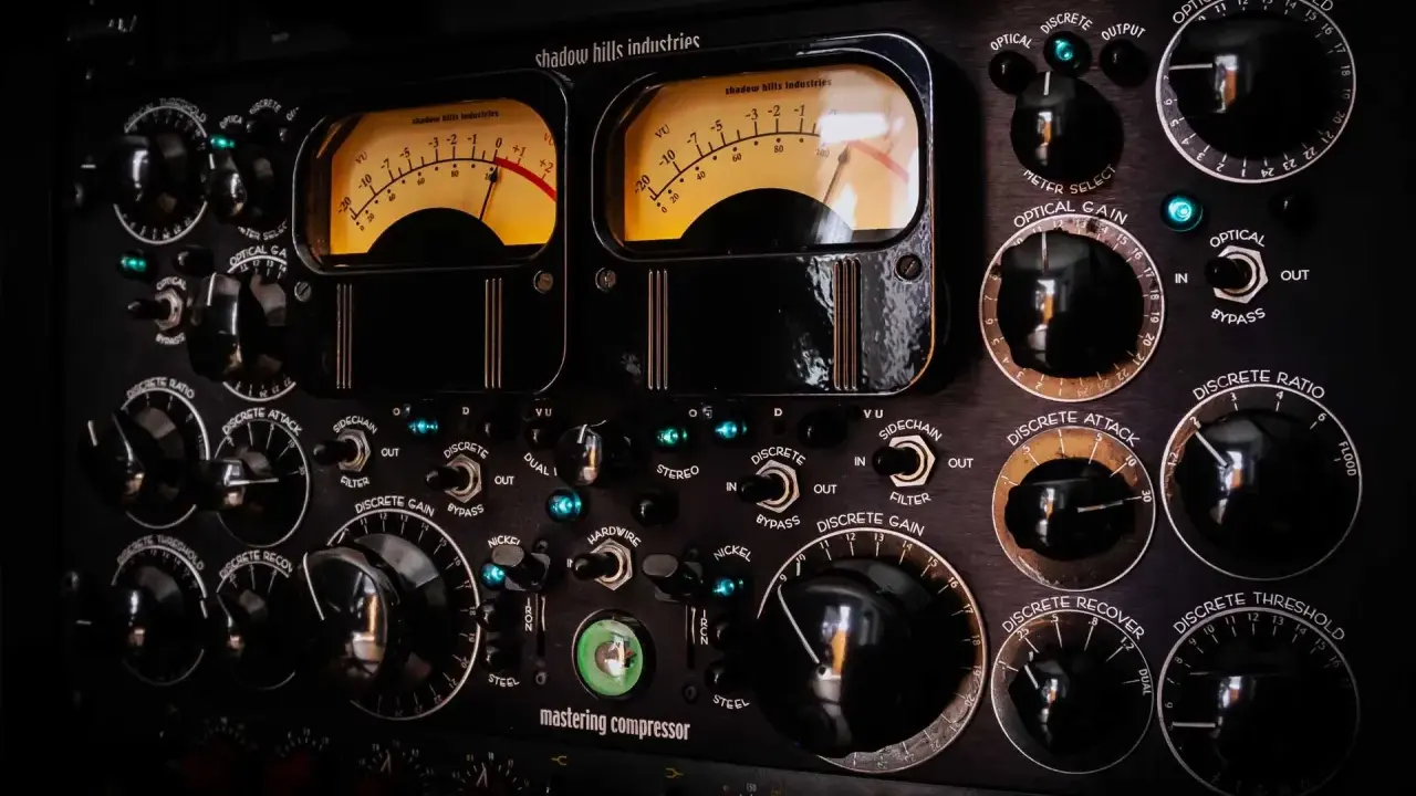 Gros plan sur un compresseur mastering avec deux VU-mètres lumineux et de nombreux boutons noirs.