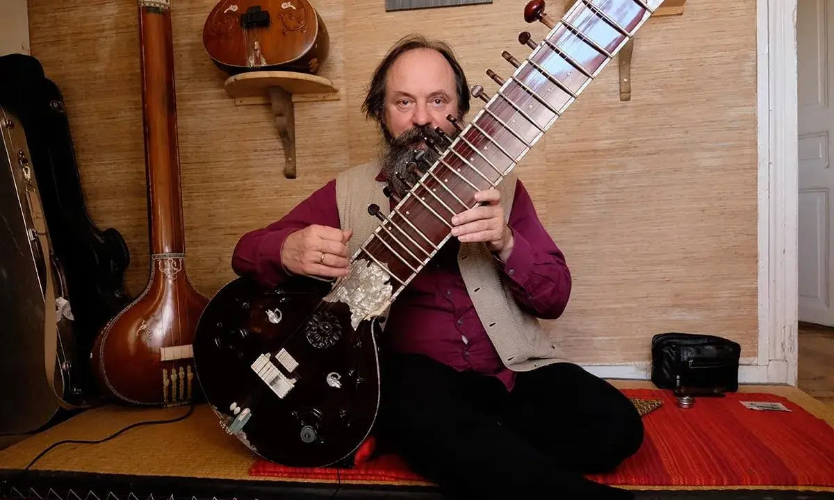 Un homme barbu joue d'un sitar. D'autres instruments de musique indienne sont visibles en arrière-plan.