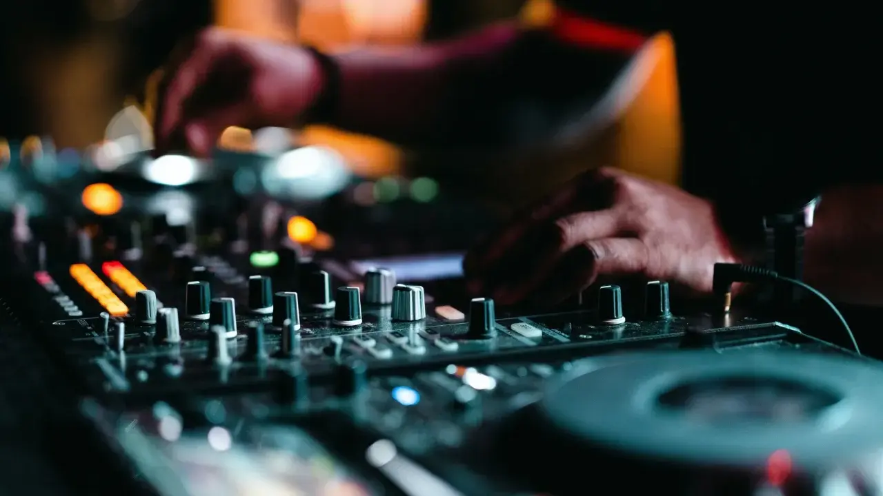 Mains d'un DJ en pleine action, manipulant les boutons pour apprendre à mixer. Lumières colorées et ambiance nocturne.