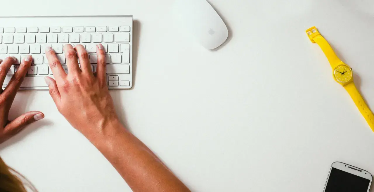 Mains sur le clavier, souris et montre jaune vif sur un bureau blanc. Un studio raccourci pour travailler.