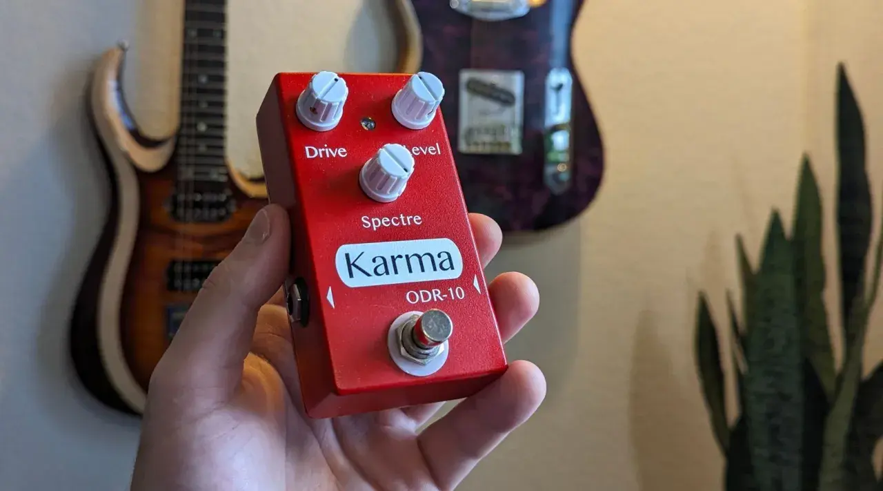 Main rouge Karma ODR-10, une pédale overdrive, tenue dans une main, avec des guitares en arrière-plan.