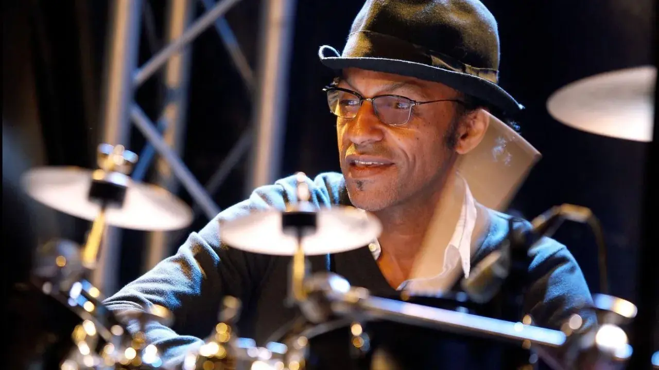 Un batteur concentré, portant un chapeau et des lunettes, joue sur scène. Il est difficile de dire combien de temps pour apprendre la batterie, mais il semble maîtriser son art.