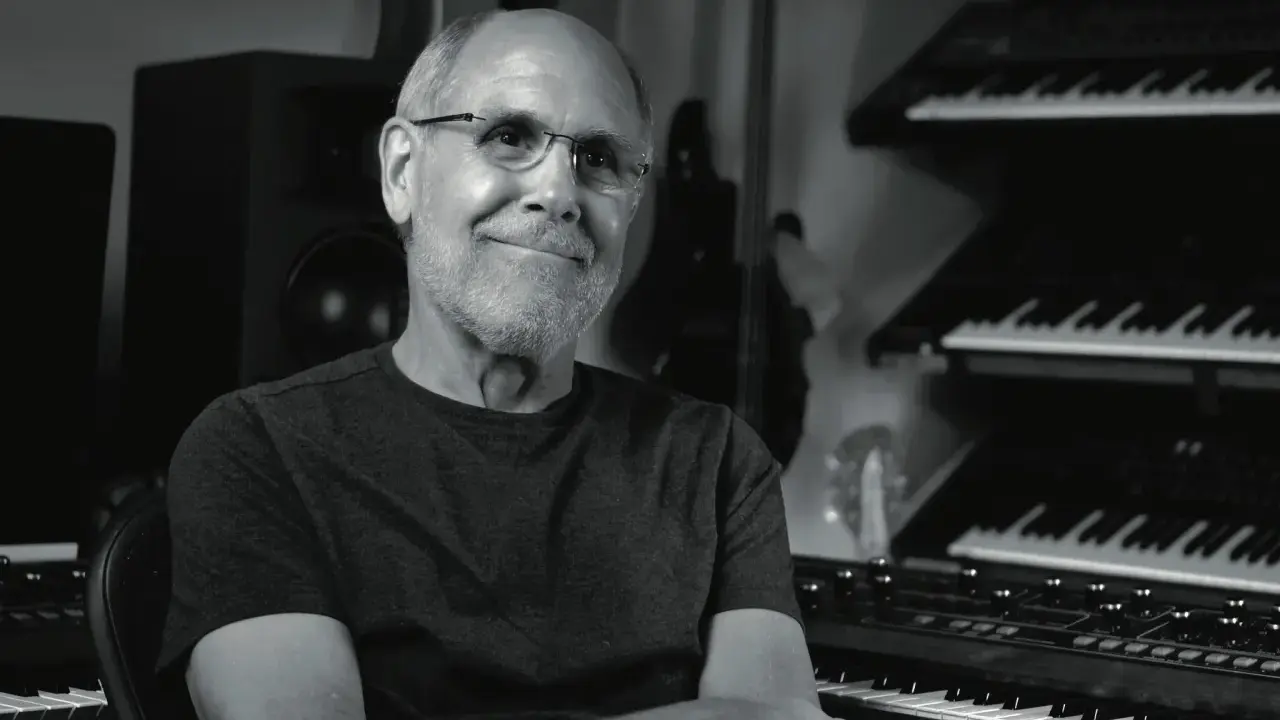 Dave Smith, souriant, dans son studio avec des synthétiseurs empilés.