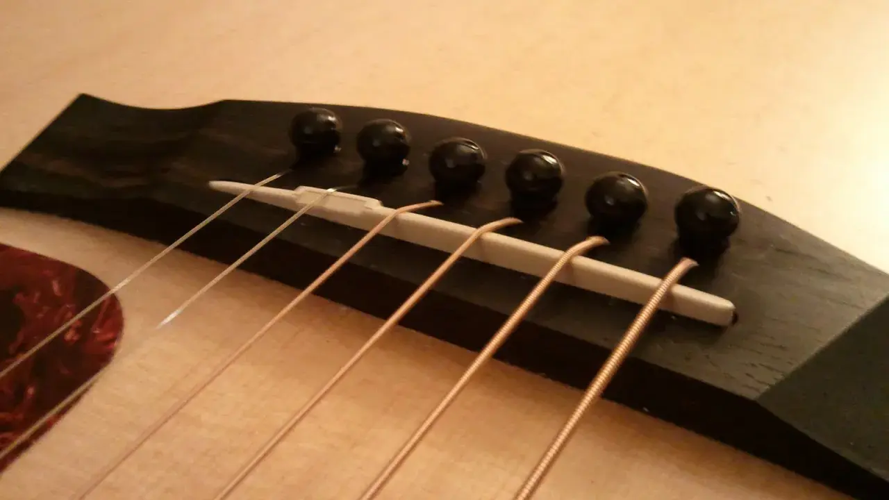 Gros plan sur le chevalet d'une guitare acoustique, avec ses six cordes prêtes à vibrer. Le bois sombre du chevalet contraste avec la pièce blanche où reposent les cordes.
