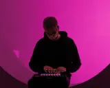 Un homme en sweat à capuche noir joue sur un contrôleur Ableton, illuminé par une lumière rose. Il est plongé dans sa création, un moment clé de la culture musicale moderne.