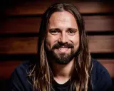 Un homme souriant aux longs cheveux bruns et à la barbe, peut-être un des compagnons d'écriture de Max Martin.