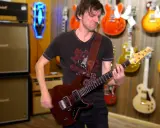 Un guitariste joue une guitare rouge, entouré de nombreuses autres guitares accrochées au mur. Un accord 5 résonne dans la pièce.