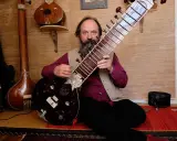 Un homme barbu joue d'un sitar. D'autres instruments de musique indienne sont visibles en arrière-plan.