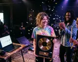 Une artiste souriante reçoit un disque d'or, entourée de musiciens applaudissant. Des confettis tombent, célébrant leur succès à créer un son unique.
