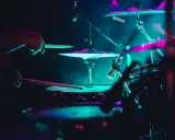 Mains d'un batteur frappant la caisse claire, avec des cymbales illuminées de néons. La **musique accompagnement batterie** crée une ambiance électrique.