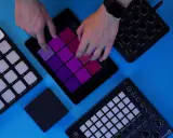 Des mains manipulent une tablette affichant des pads colorés, entourée de contrôleurs midi. Création musicale en cours.
