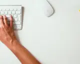 Mains sur le clavier, souris et montre jaune vif sur un bureau blanc. Un studio raccourci pour travailler.
