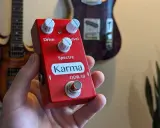 Main rouge Karma ODR-10, une pédale overdrive, tenue dans une main, avec des guitares en arrière-plan.