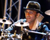 Un batteur concentré, portant un chapeau et des lunettes, joue sur scène. Il est difficile de dire combien de temps pour apprendre la batterie, mais il semble maîtriser son art.