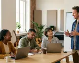 Un formateur anime une session de formation MAO CPF. Trois apprenants sont attentifs autour d'une table, certains sur ordinateur, d'autres prenant des notes.
