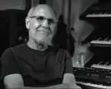 Dave Smith, souriant, dans son studio avec des synthétiseurs empilés.