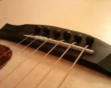 Gros plan sur le chevalet d'une guitare acoustique, avec ses six cordes prêtes à vibrer. Le bois sombre du chevalet contraste avec la pièce blanche où reposent les cordes.