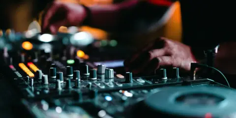 Mains d'un DJ en pleine action, manipulant les boutons pour apprendre à mixer. Lumières colorées et ambiance nocturne.