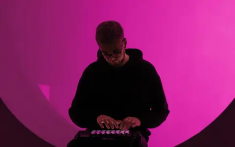 Un homme en sweat à capuche noir joue sur un contrôleur Ableton, illuminé par une lumière rose. Il est plongé dans sa création, un moment clé de la culture musicale moderne.
