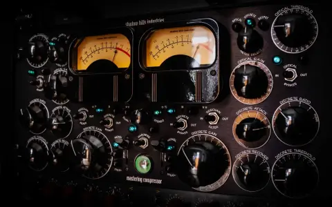 Gros plan sur un compresseur mastering avec deux VU-mètres lumineux et de nombreux boutons noirs.