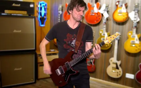 Un guitariste joue une guitare rouge, entouré de nombreuses autres guitares accrochées au mur. Un accord 5 résonne dans la pièce.