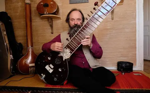 Un homme barbu joue d'un sitar. D'autres instruments de musique indienne sont visibles en arrière-plan.