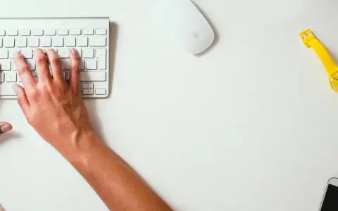 Mains sur le clavier, souris et montre jaune vif sur un bureau blanc. Un studio raccourci pour travailler.