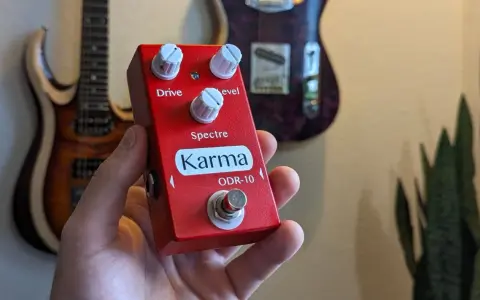 Main rouge Karma ODR-10, une pédale overdrive, tenue dans une main, avec des guitares en arrière-plan.