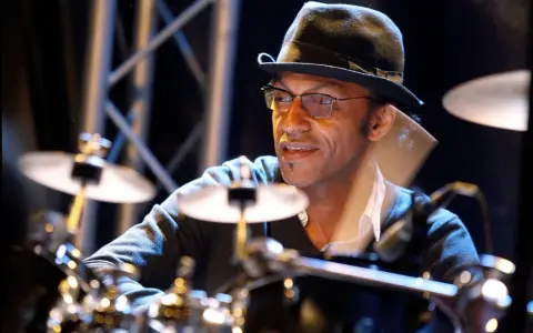 Un batteur concentré, portant un chapeau et des lunettes, joue sur scène. Il est difficile de dire combien de temps pour apprendre la batterie, mais il semble maîtriser son art.