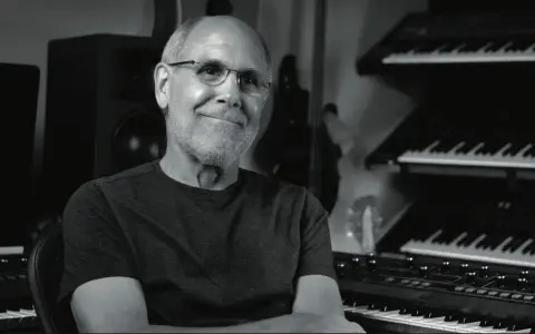Dave Smith, souriant, dans son studio avec des synthétiseurs empilés.