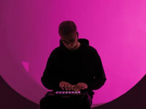 Un homme en sweat à capuche noir joue sur un contrôleur Ableton, illuminé par une lumière rose. Il est plongé dans sa création, un moment clé de la culture musicale moderne.