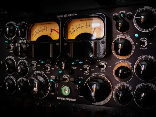 Gros plan sur un compresseur mastering avec deux VU-mètres lumineux et de nombreux boutons noirs.