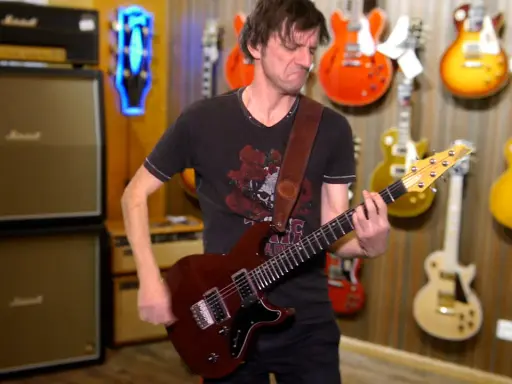 Un guitariste joue une guitare rouge, entouré de nombreuses autres guitares accrochées au mur. Un accord 5 résonne dans la pièce.