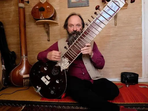 Un homme barbu joue d'un sitar. D'autres instruments de musique indienne sont visibles en arrière-plan.