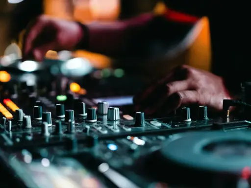 Mains d'un DJ en pleine action, manipulant les boutons pour apprendre à mixer. Lumières colorées et ambiance nocturne.