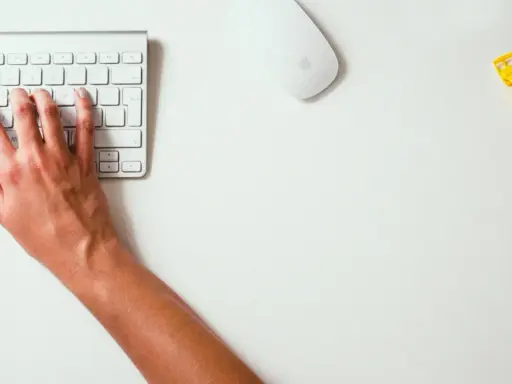 Mains sur le clavier, souris et montre jaune vif sur un bureau blanc. Un studio raccourci pour travailler.