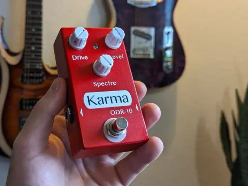 Main rouge Karma ODR-10, une pédale overdrive, tenue dans une main, avec des guitares en arrière-plan.
