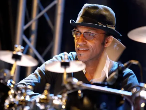 Un batteur concentré, portant un chapeau et des lunettes, joue sur scène. Il est difficile de dire combien de temps pour apprendre la batterie, mais il semble maîtriser son art.