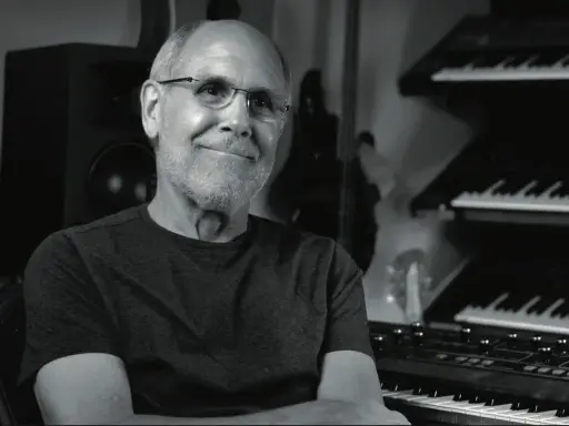Dave Smith, souriant, dans son studio avec des synthétiseurs empilés.
