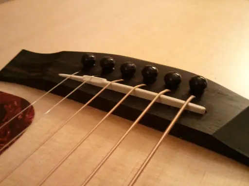 Gros plan sur le chevalet d'une guitare acoustique, avec ses six cordes prêtes à vibrer. Le bois sombre du chevalet contraste avec la pièce blanche où reposent les cordes.