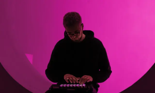 Un homme en sweat à capuche noir joue sur un contrôleur Ableton, illuminé par une lumière rose. Il est plongé dans sa création, un moment clé de la culture musicale moderne.