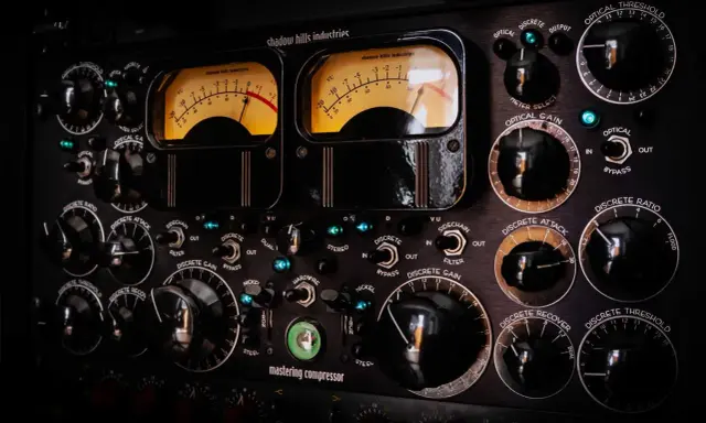 Gros plan sur un compresseur mastering avec deux VU-mètres lumineux et de nombreux boutons noirs.
