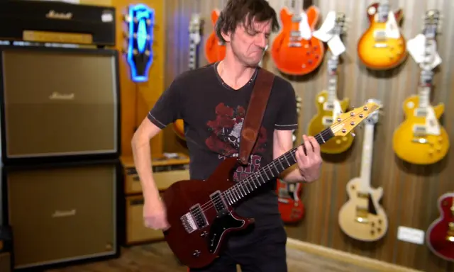 Un guitariste joue une guitare rouge, entouré de nombreuses autres guitares accrochées au mur. Un accord 5 résonne dans la pièce.
