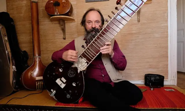 Un homme barbu joue d'un sitar. D'autres instruments de musique indienne sont visibles en arrière-plan.