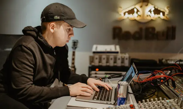 Un jeune homme travaille sur un ordinateur portable dans un studio d'enregistrement, avec le logo Red Bull en arrière-plan.