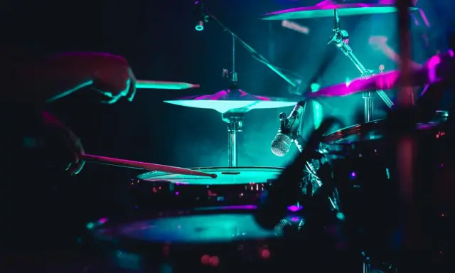Mains d'un batteur frappant la caisse claire, avec des cymbales illuminées de néons. La **musique accompagnement batterie** crée une ambiance électrique.