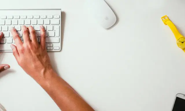 Mains sur le clavier, souris et montre jaune vif sur un bureau blanc. Un studio raccourci pour travailler.