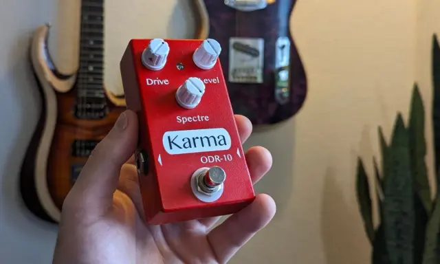 Main rouge Karma ODR-10, une pédale overdrive, tenue dans une main, avec des guitares en arrière-plan.