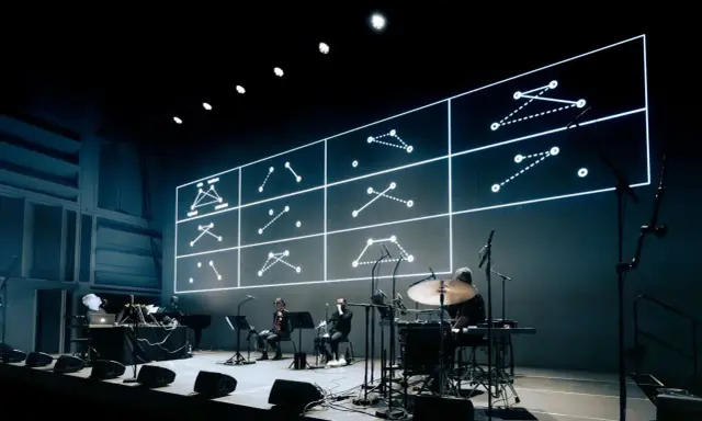 Sur scène, des musiciens jouent devant un écran projetant des schémas lumineux. Une performance pour comment se faire remarquer.